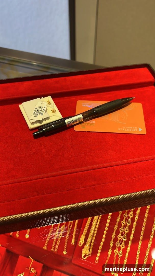 A close-up view of a red velvet-lined display case, likely in a jewelry store. On the velvet surface, a black pen rests on top of an orange card that says "LIFESTYLE REWARD". To the left of the pen is a small white tag attached to a pair of gold earrings. Below the velvet surface, a display of various gold necklaces and bracelets is visible through a glass pane.