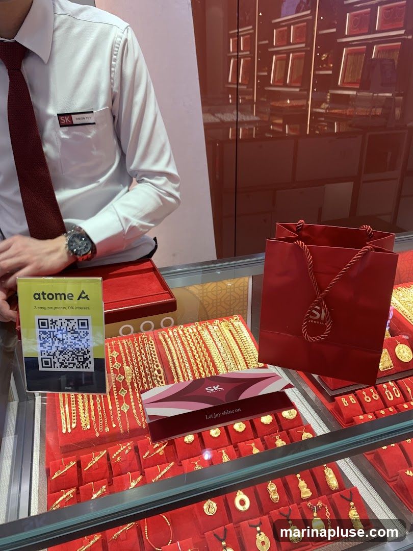 A display case filled with gold jewelry, including bracelets and pendants. A person in a white shirt and red tie stands behind the counter, wearing a name tag that reads 'SK DEON TEY'. A red shopping bag with the SK Jewellery logo is visible. A sign for 'atome A' with a QR code is also present.