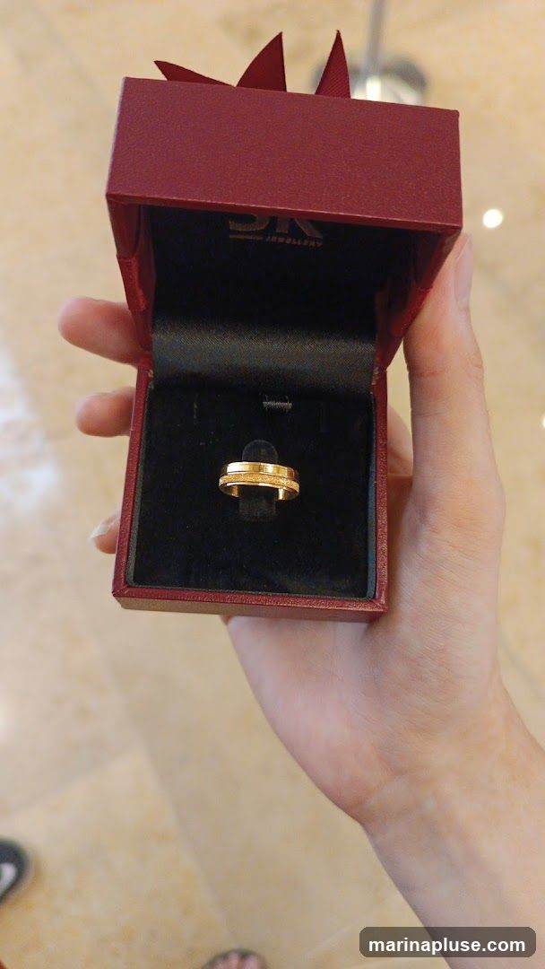 A hand holds open a dark red jewelry box revealing a gold ring with a textured band. The inside of the box has the logo "DK JEWELLERY" printed on it.