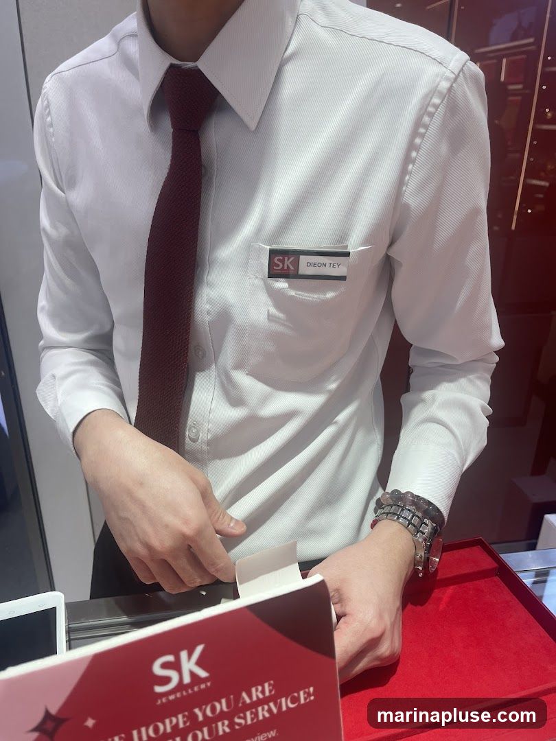 A person wearing a white collared shirt and a maroon tie. A name tag on the shirt reads "SK JEWELLERY DION TEY". The person is standing behind a counter with a red velvet surface. A small white box is open in front of them. A red card with "SK JEWELLERY" and text about service is visible in the foreground.