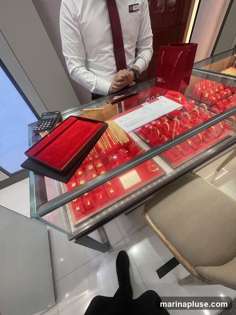 A display case filled with gold jewelry, including necklaces, bracelets, and rings, is shown in a jewelry store. A salesperson in a white shirt and red tie stands behind the counter. A calculator and a red velvet jewelry box are also visible. A red shopping bag with the logo "SK" is on the counter.