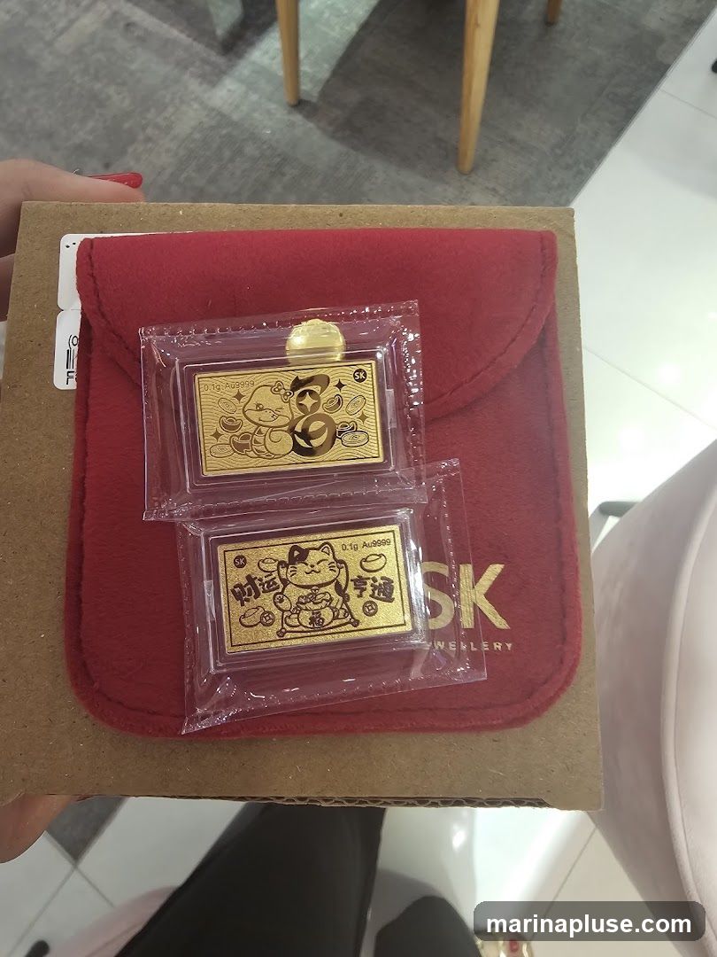 Two small gold bars, each 0.1g Au9999, are displayed on a red pouch. One bar features a character resembling Hello Kitty surrounded by coins and Chinese characters. The other bar depicts a Maneki-neko (beckoning cat) with Chinese characters. The red pouch has the text "SK JEWELLERY" visible. The items are placed on a cardboard box, and a person's hand is holding the box.