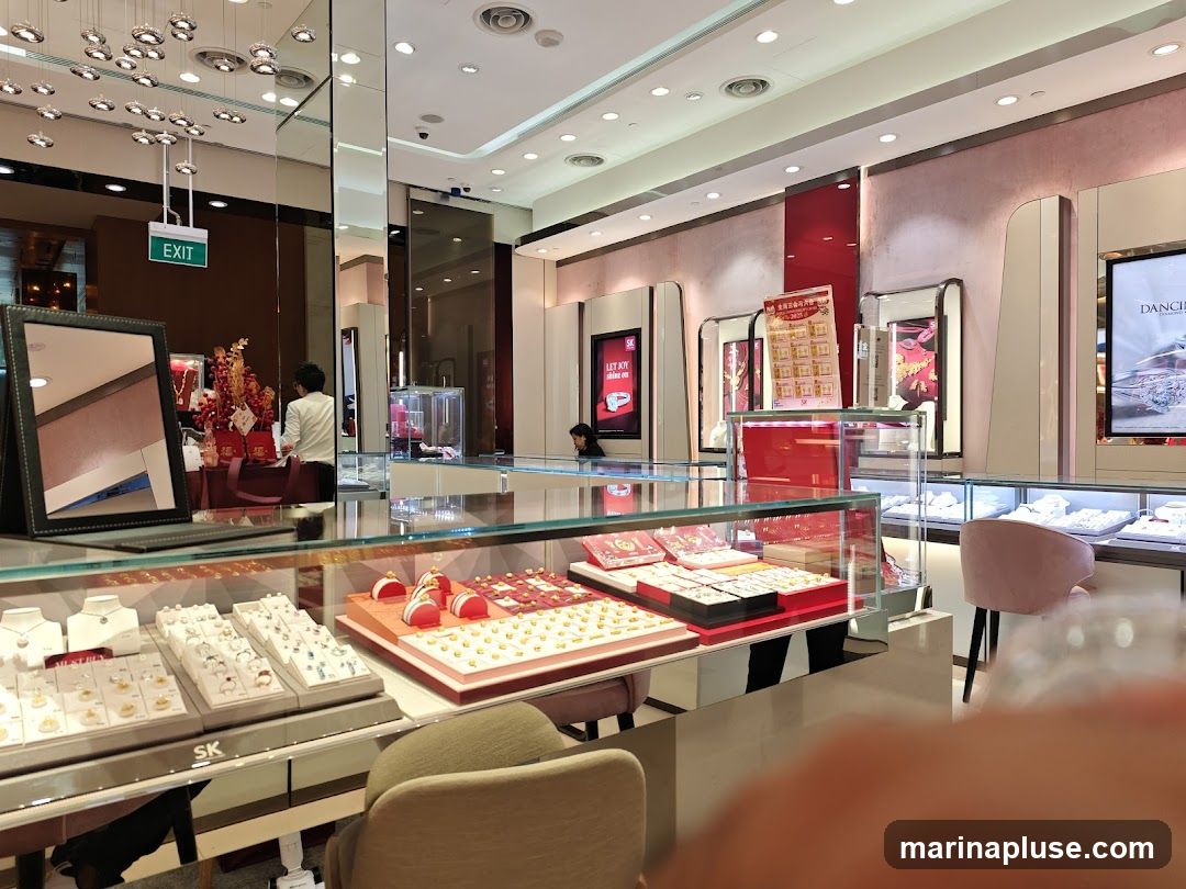 A well-lit jewelry store interior with display cases filled with necklaces, rings, and gold ornaments. Several customers and staff members are visible. A large screen displays an advertisement for DANCING.