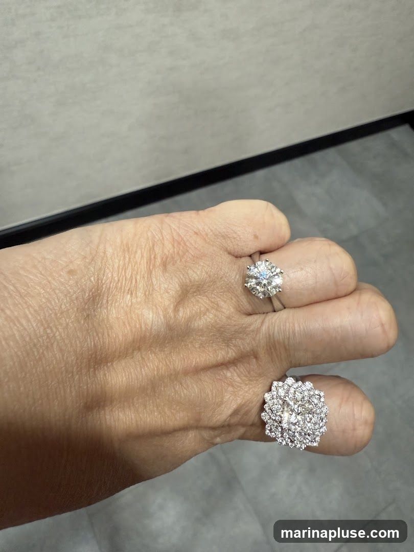 A close-up shot of a hand wearing two diamond rings. One ring is on the ring finger and features a large, round-cut diamond. The other ring is on the middle finger and is a cluster ring with many smaller diamonds arranged in a floral pattern. The background is a textured wall and a grey floor.