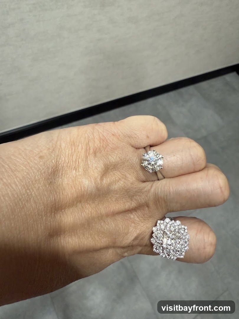 A close-up shot of a hand wearing two diamond rings. One ring is on the ring finger and features a large, round-cut diamond. The other ring is on the middle finger and is a cluster ring with many smaller diamonds arranged in a floral pattern. The background is a textured wall and a grey floor.