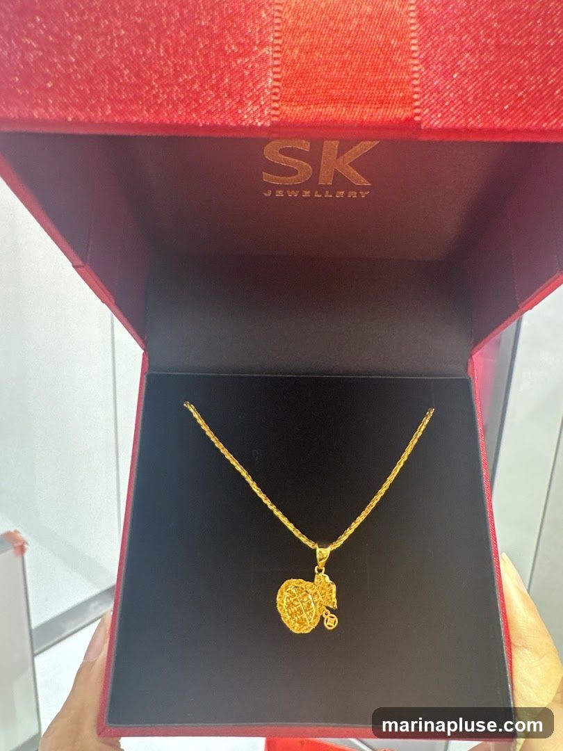 A gold necklace with a pendant shaped like a traditional Chinese money bag is displayed inside a red jewelry box. The brand name 'SK JEWELLERY' is visible on the inside lid of the box.