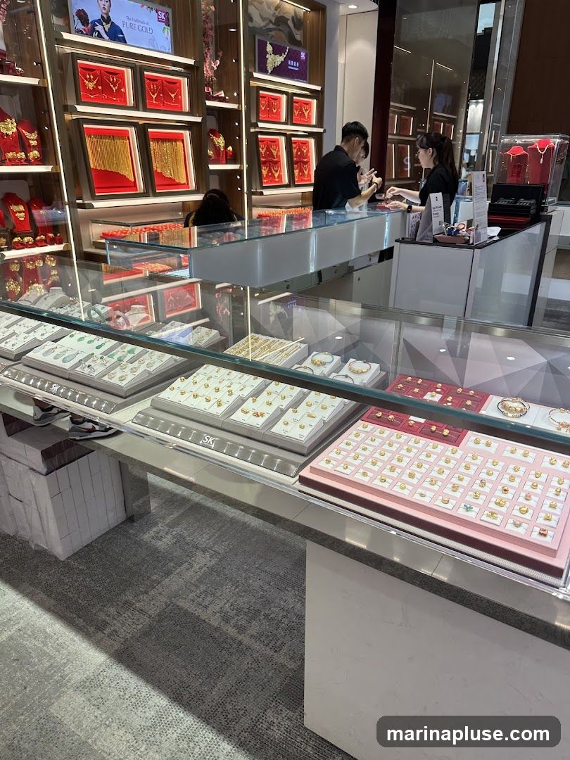 A well-lit jewelry store interior with displays of gold necklaces, bracelets, and rings. Two staff members are visible behind a counter assisting a customer. The shelves and display cases are filled with various gold jewelry items, some presented in red velvet boxes. A sign above the display reads "SK PURE GOLD".