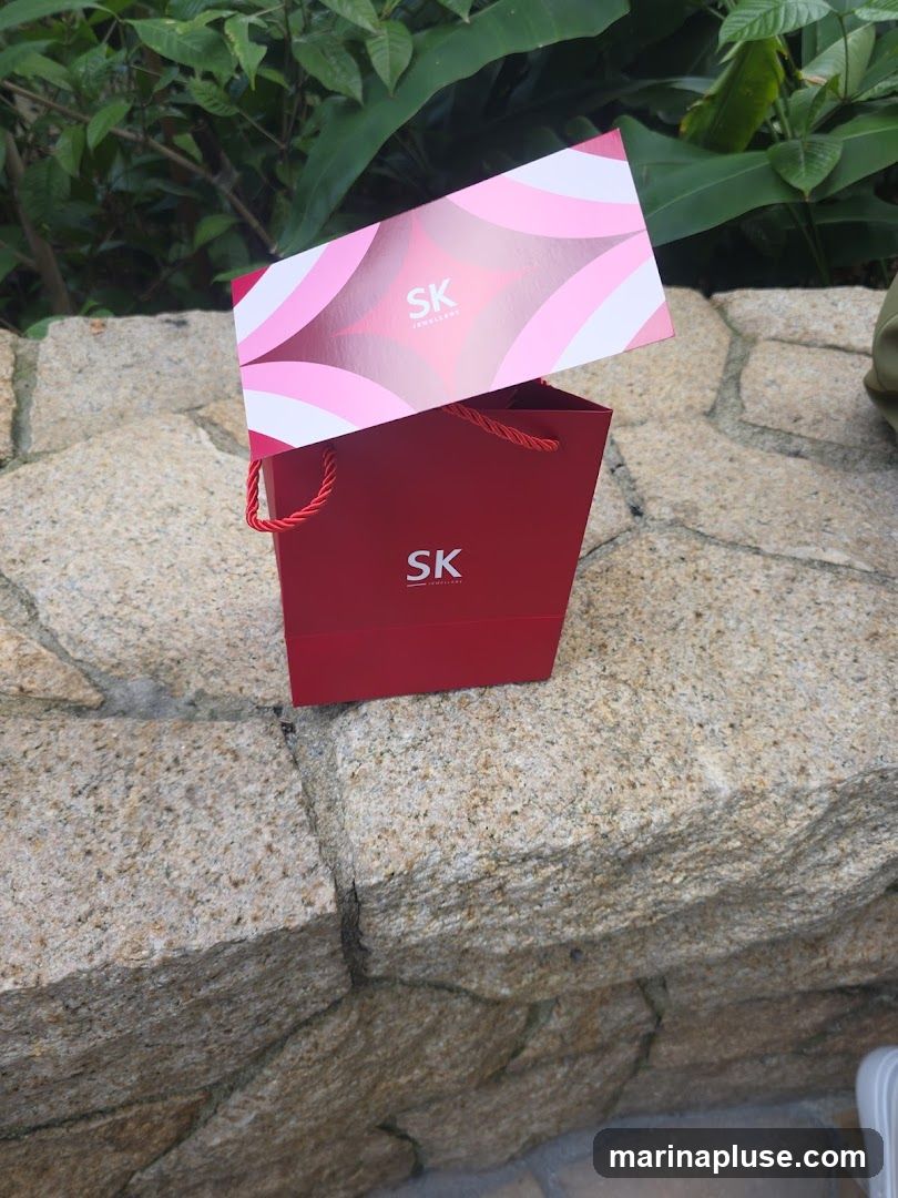 A red gift bag with a card on top, both featuring the SK Jewellery logo. The bag has red rope handles. The background consists of green foliage and a stone wall.