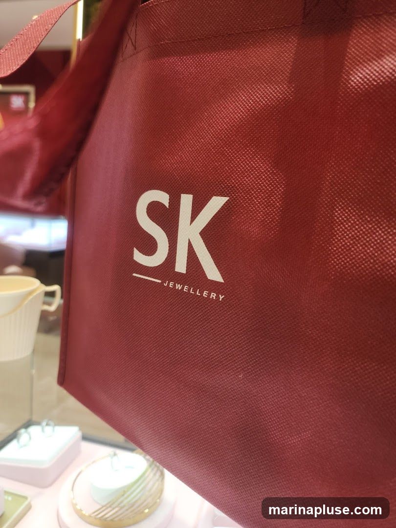 A close-up of a red reusable bag with the white text "SK JEWELLERY" printed on it. The bag is held up, obscuring most of the background, but a jewelry display case with rings and other items is visible below.