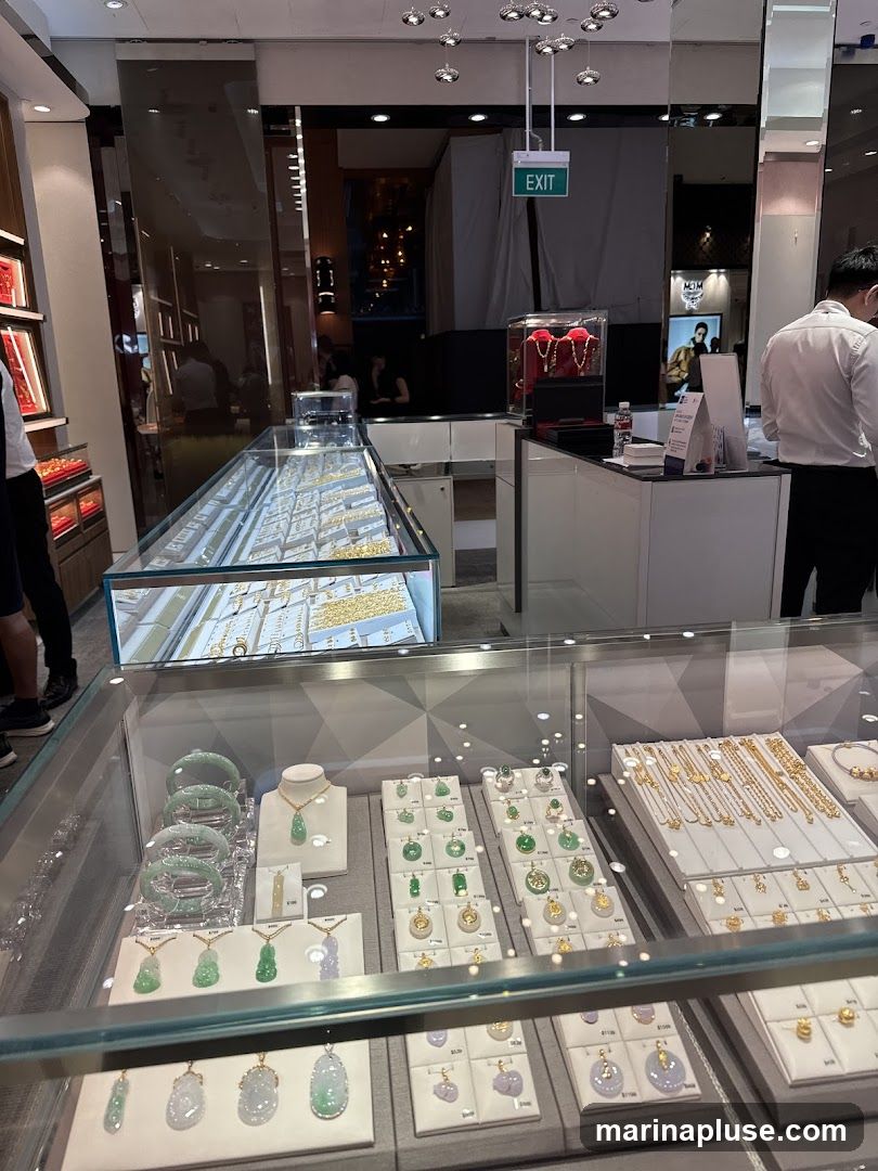 A well-lit jewelry store interior with multiple display cases filled with gold and jade jewelry. The cases showcase necklaces, bracelets, pendants, and rings. In the background, staff members can be seen attending to customers and behind a counter. An "EXIT" sign is visible overhead.