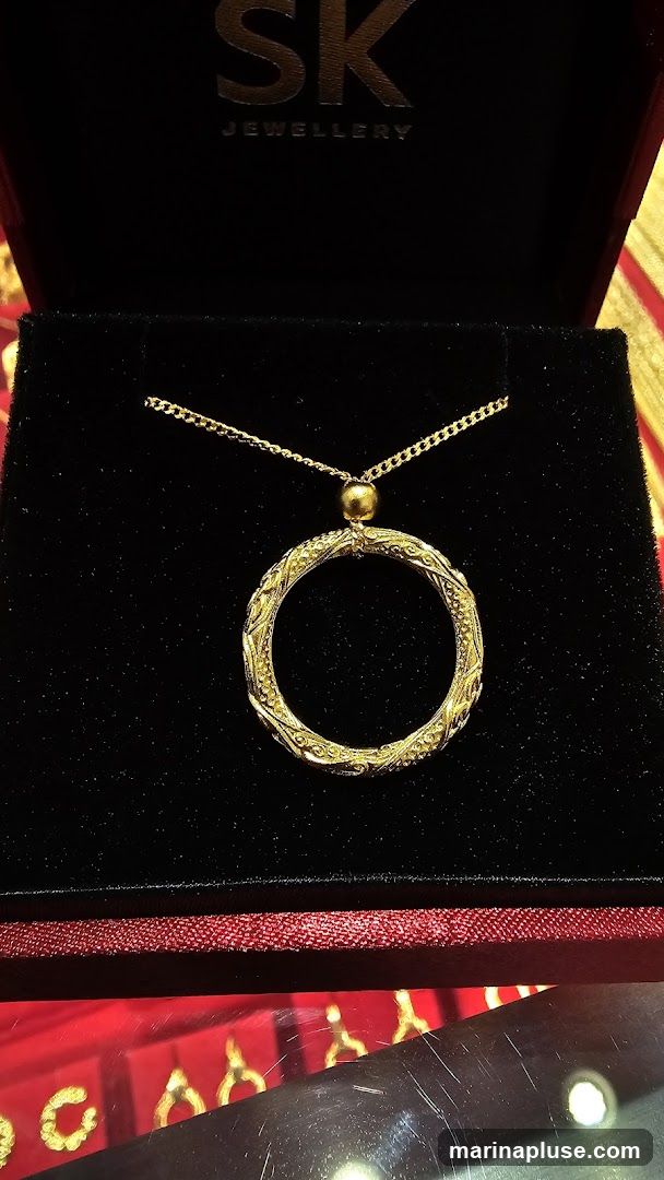A close-up shot of a gold pendant necklace displayed in a black velvet jewelry box. The pendant is circular with intricate, ornate detailing. The box has the SK Jewellery logo embossed in silver on the inside lid.