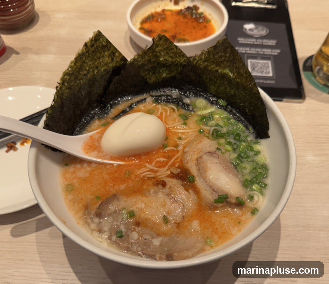 A close-up shot of a bowl of ramen with a creamy broth, topped with nori seaweed, a soft-boiled egg, chashu pork, and chopped green onions. A spoon is in the bowl, lifting some noodles. In the background, a small dish of sauce and a menu board with a QR code are visible.