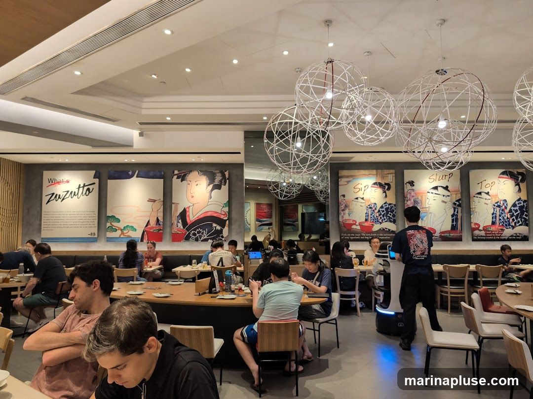 The image shows the interior of a restaurant with several patrons seated at tables. Large, artistic posters featuring Japanese-style illustrations and text are displayed on the walls. The posters have titles like "What's Zuzutto", "Sip", and "Top". Modern, spherical light fixtures hang from the ceiling. A robot server is visible in the middle of the dining area. The overall ambiance appears to be casual and modern.
