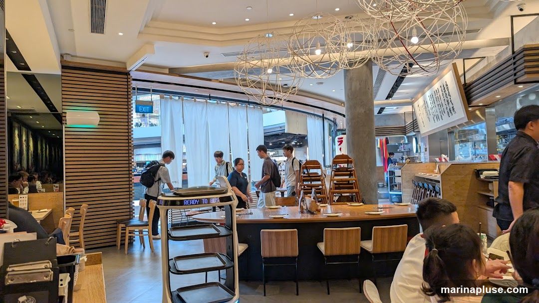 The image shows the interior of a restaurant with several customers and staff members. A robotic food delivery cart is visible in the foreground. The restaurant has a modern design with wooden accents and decorative lighting fixtures. A large menu board is displayed on the wall.