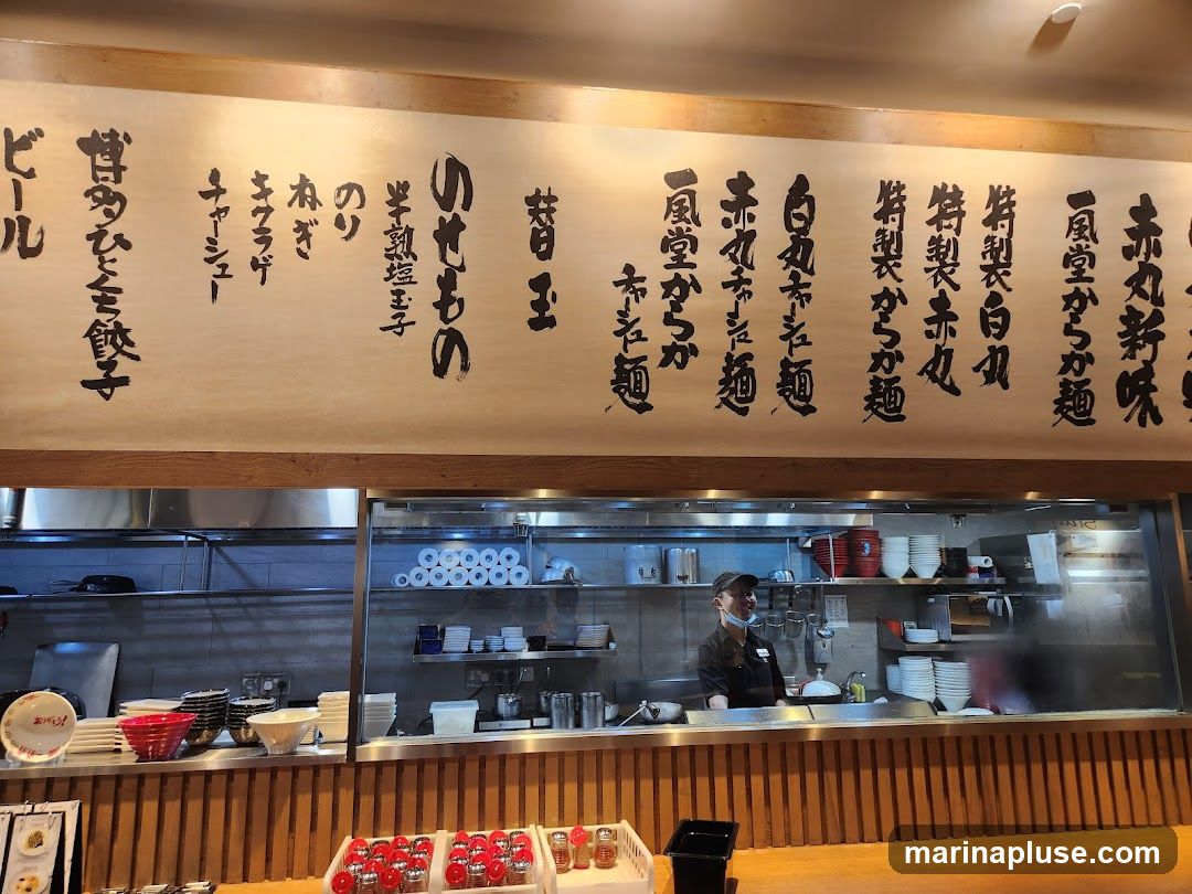 A view of a restaurant kitchen with a large handwritten menu board above. The menu features various ramen dishes and side items. A staff member is visible working in the kitchen. Bowls and other kitchenware are neatly arranged on shelves and counters.