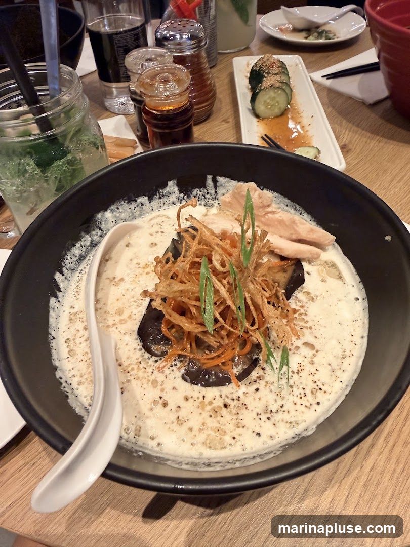 A close-up shot of a bowl of ramen. The broth is creamy and white, topped with shredded fried onions, sliced chicken, and pieces of eggplant. A white spoon rests on the side of the bowl. In the background, there are drinks, condiments, and a plate of sliced cucumbers.
