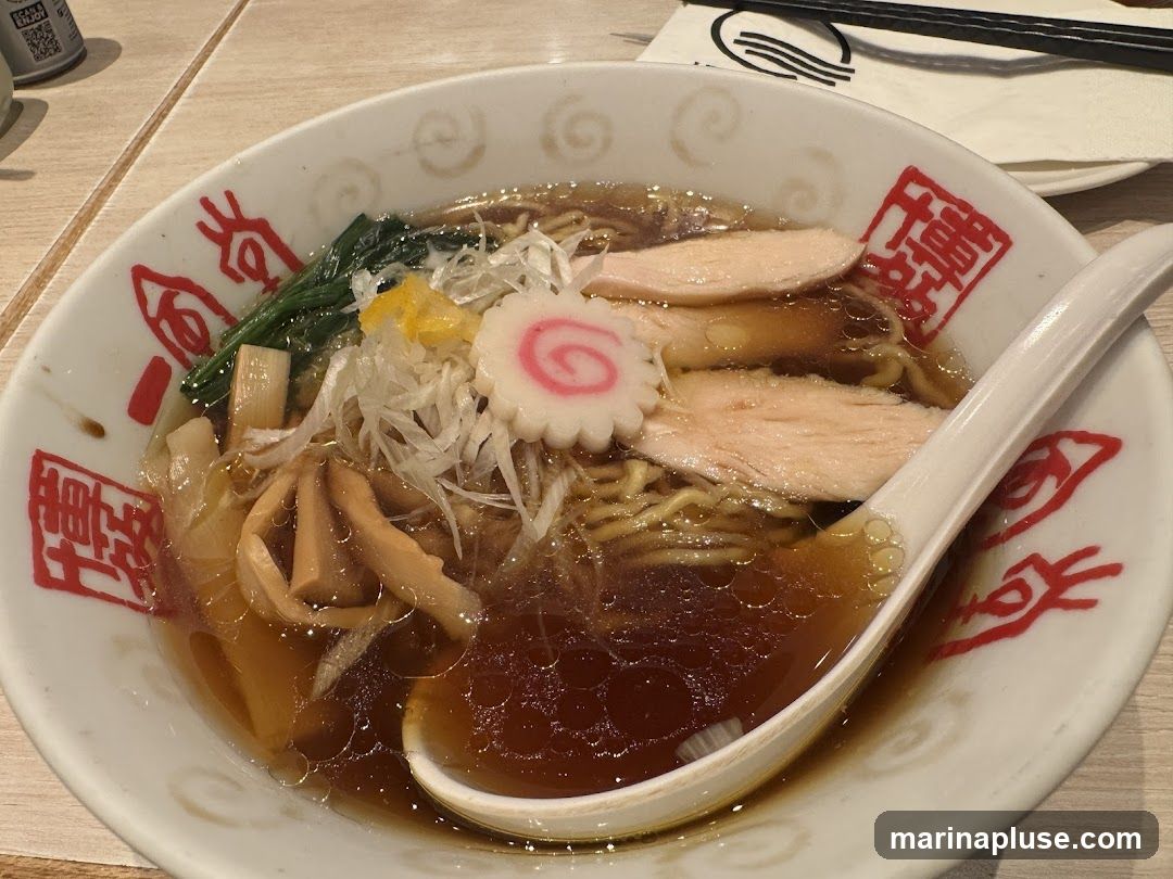 A bowl of ramen with chicken slices, narutomaki (fish cake), bamboo shoots, green onions, and spinach in a savory broth. The bowl has red Japanese characters and a white spoon rests inside.