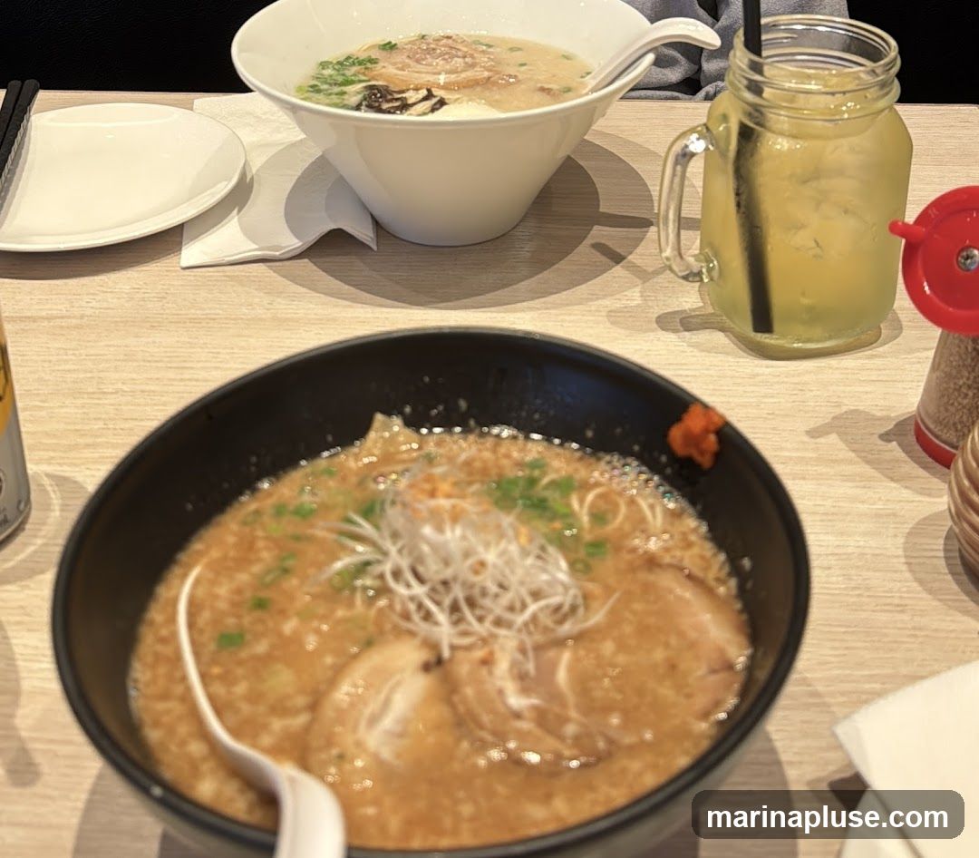 Two bowls of ramen and a glass of yellow drink on a wooden table. One bowl is black and contains ramen with pork slices and green onions. The other bowl is white and contains ramen with pork slices and green onions. A glass jar with a handle contains a yellow beverage with ice and a black straw. A white napkin and a white plate are also visible.
