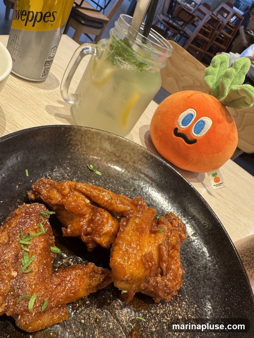 A close-up shot of several fried chicken wings on a dark plate, garnished with chopped green herbs. To the left, a can of Schweppes is visible. In the background, a glass jar filled with a lemonade-like drink with mint and lemon slices is present, along with a small, orange plush toy resembling a carrot. The setting appears to be a restaurant table.