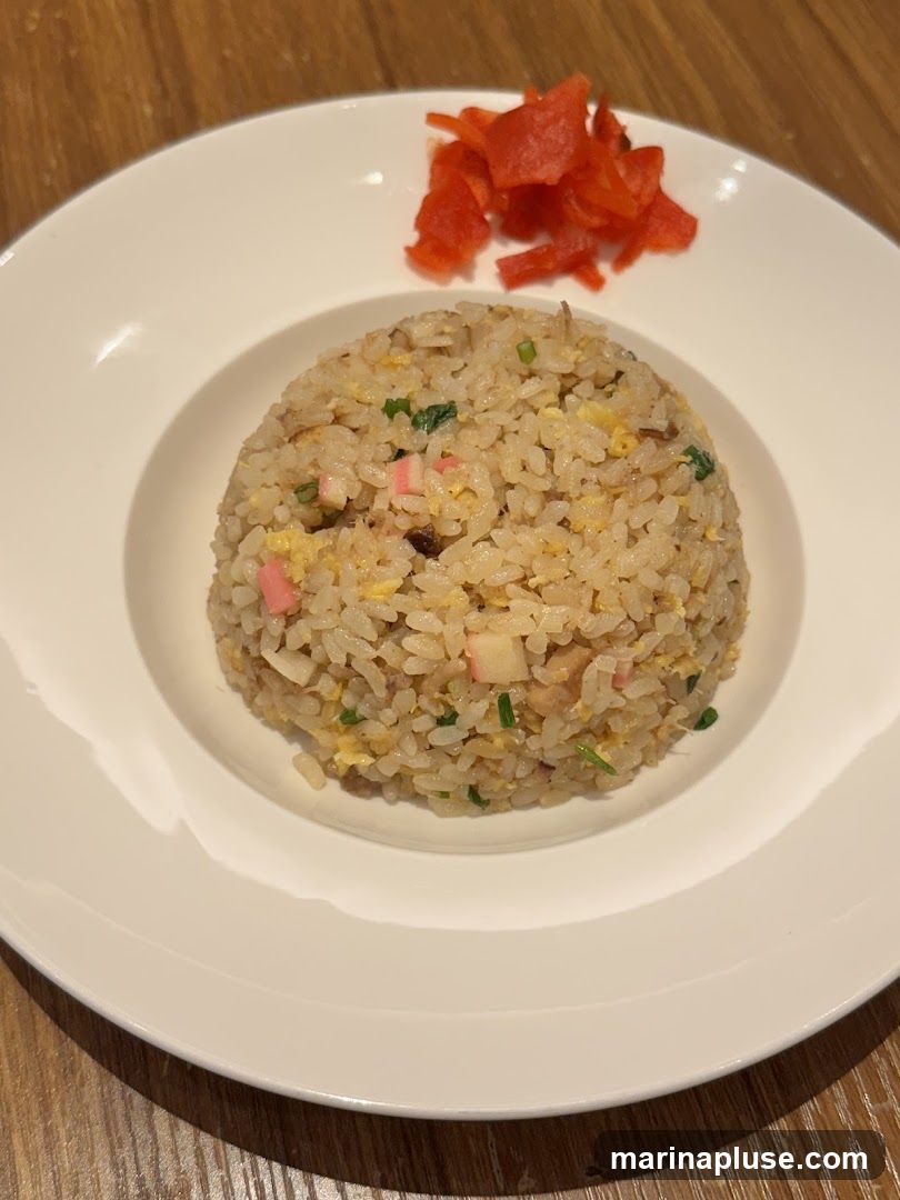 A close-up shot of a serving of fried rice on a white plate. The fried rice is a mound in the center of the plate and appears to contain small pieces of egg, green onions, and possibly some type of seafood or meat. To the top right of the fried rice, there is a small pile of bright red pickled ginger. The plate is on a wooden surface.