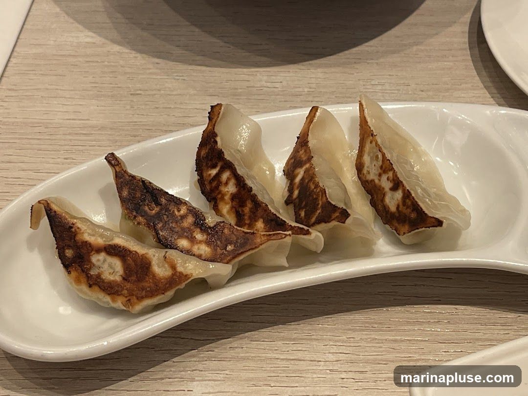 A white, curved dish holds five pan-fried dumplings. The dumplings have a golden-brown, crispy bottom and a white, steamed top. They are arranged in a row, slightly overlapping. The dish is placed on a light-colored wooden table.