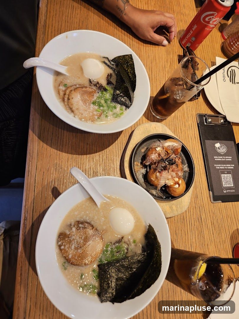 Two bowls of ramen, a small plate of takoyaki, and two drinks are arranged on a wooden table. A can of Coca-Cola is also visible. A menu is clipped to a board on the right side of the image.