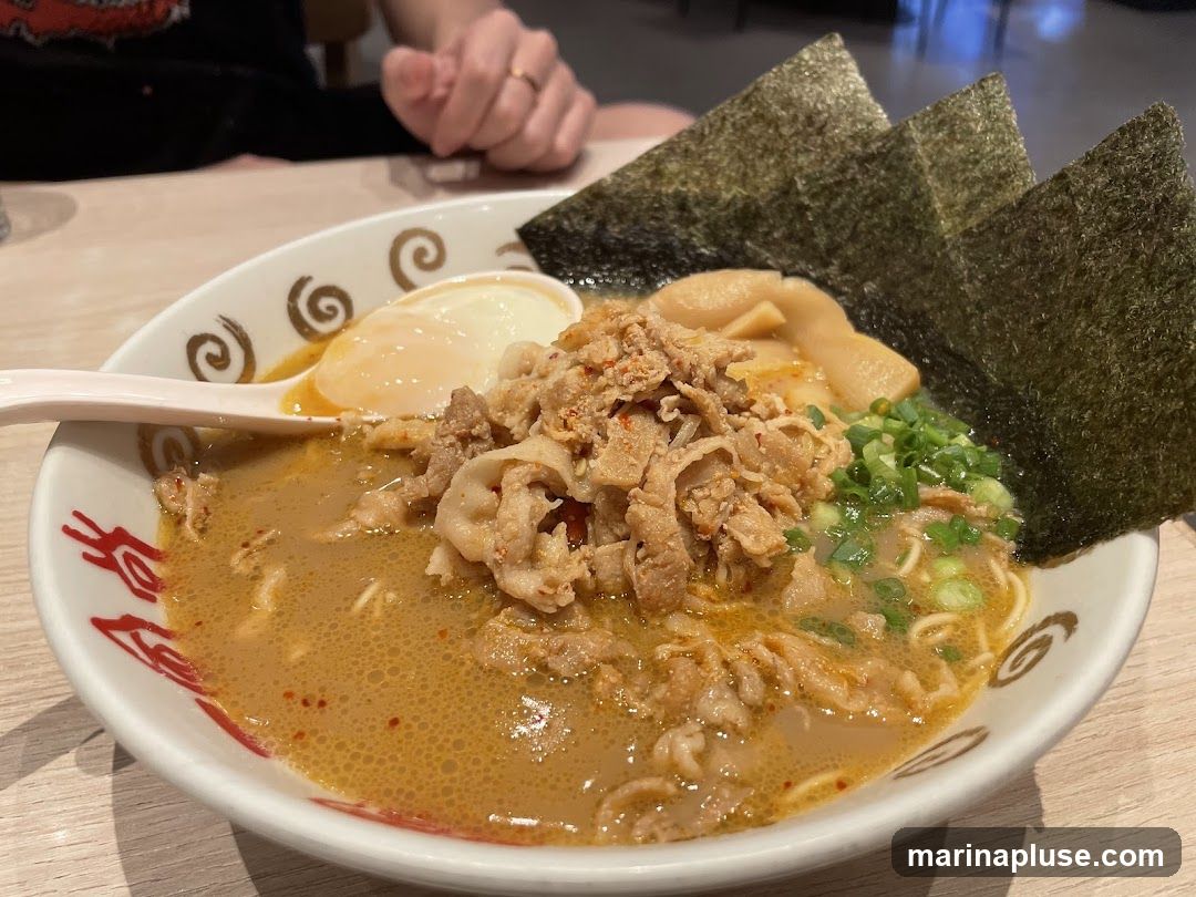 A close-up shot of a bowl of ramen. The ramen is topped with thinly sliced pork, a soft-boiled egg, green onions, bamboo shoots, and nori seaweed. The bowl has a decorative pattern around the rim.