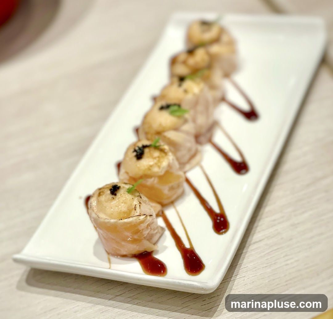 A white rectangular plate holds six salmon rolls, arranged in a diagonal line. Each roll is topped with a small sprig of green garnish and a few black beads, possibly roe. The rolls are drizzled with a dark sauce, creating abstract patterns on the plate. The background is blurred, suggesting an indoor dining setting.