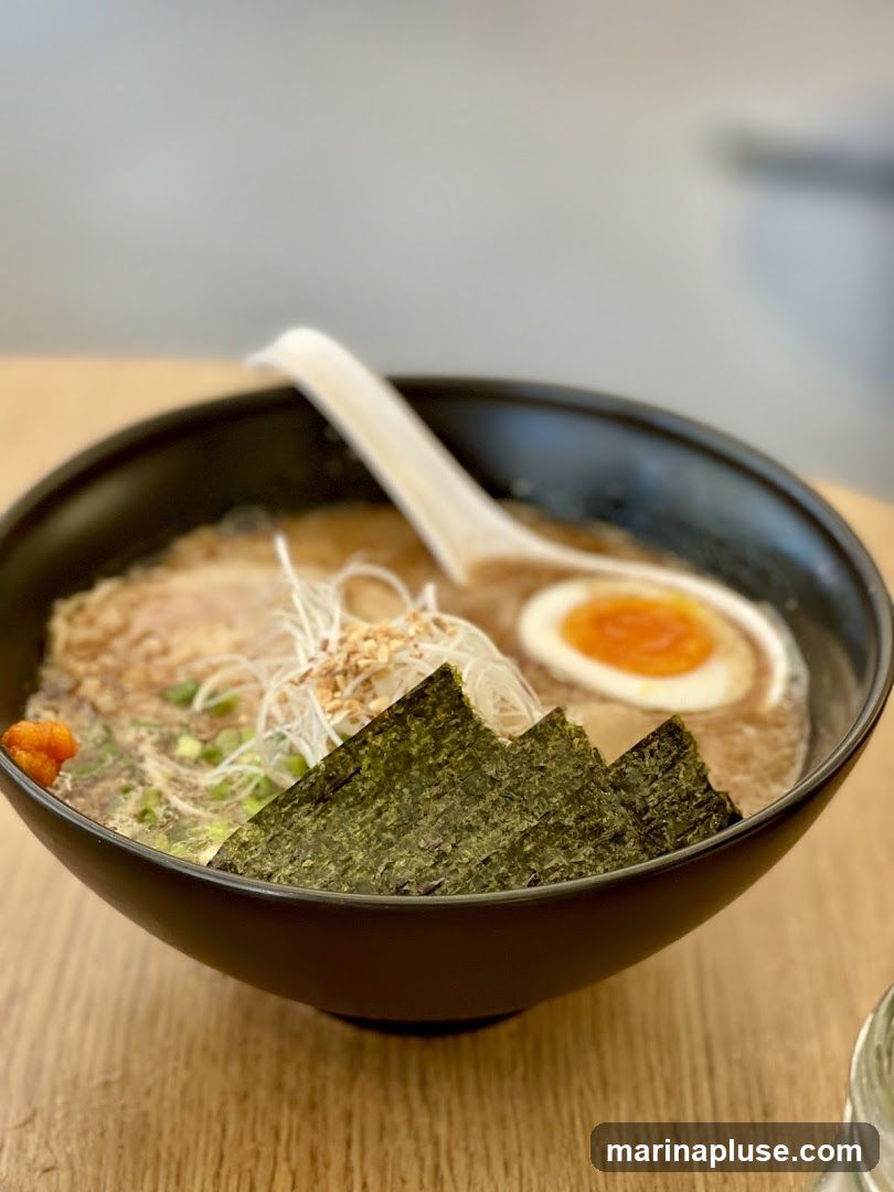A close-up shot of a ramen bowl filled with broth, noodles, sliced pork, a soft-boiled egg, nori seaweed, and green onions. The bowl is black and sits on a wooden table. A white spoon rests in the bowl.