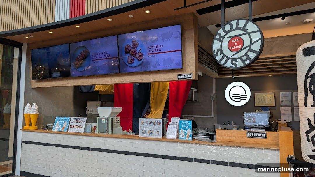 A sushi restaurant counter with digital menu boards displaying food items like Unagi & Ikura Avocado Puree and Pork Belly Jalapeno. A circular sign above reads "ROLL MAKI & TATAMI SUSHI". There are also soft serve ice cream cones displayed, and a sign that says "ORDER HERE".