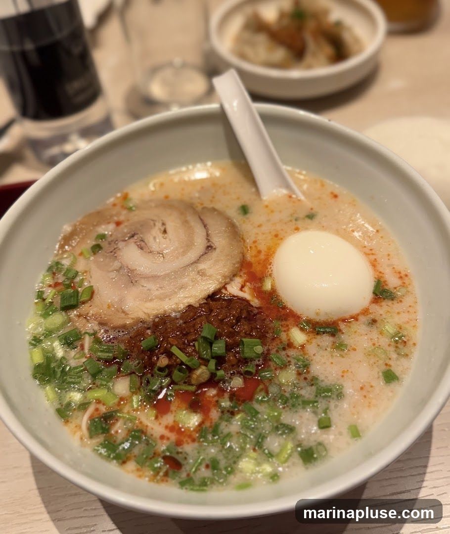 A close-up shot of a bowl of ramen. The ramen has a creamy broth, topped with a slice of chashu pork, a soft-boiled egg, chopped green onions, and a dollop of chili paste. In the background, out of focus, are a glass of water, a bottle, and a small bowl of dumplings.