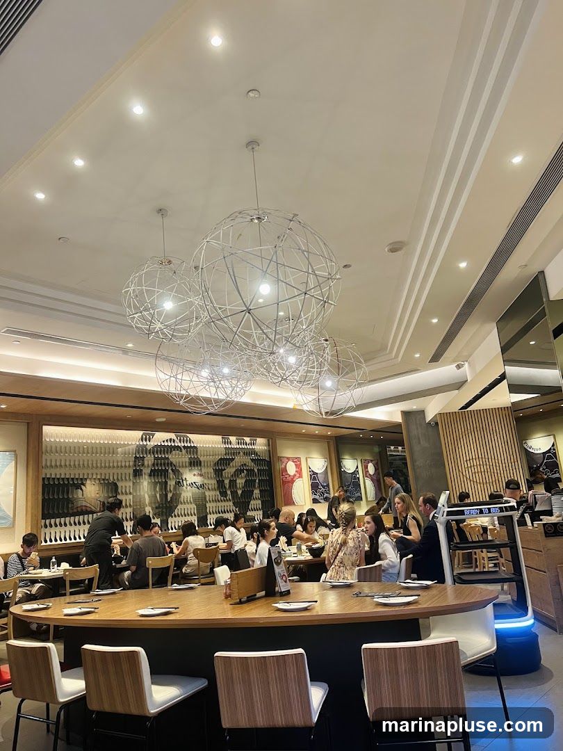The interior of a busy restaurant with customers seated at tables and a large counter. Large, spherical wire chandeliers hang from the ceiling. A decorative mural is visible on the back wall. A robot is present near a service station.