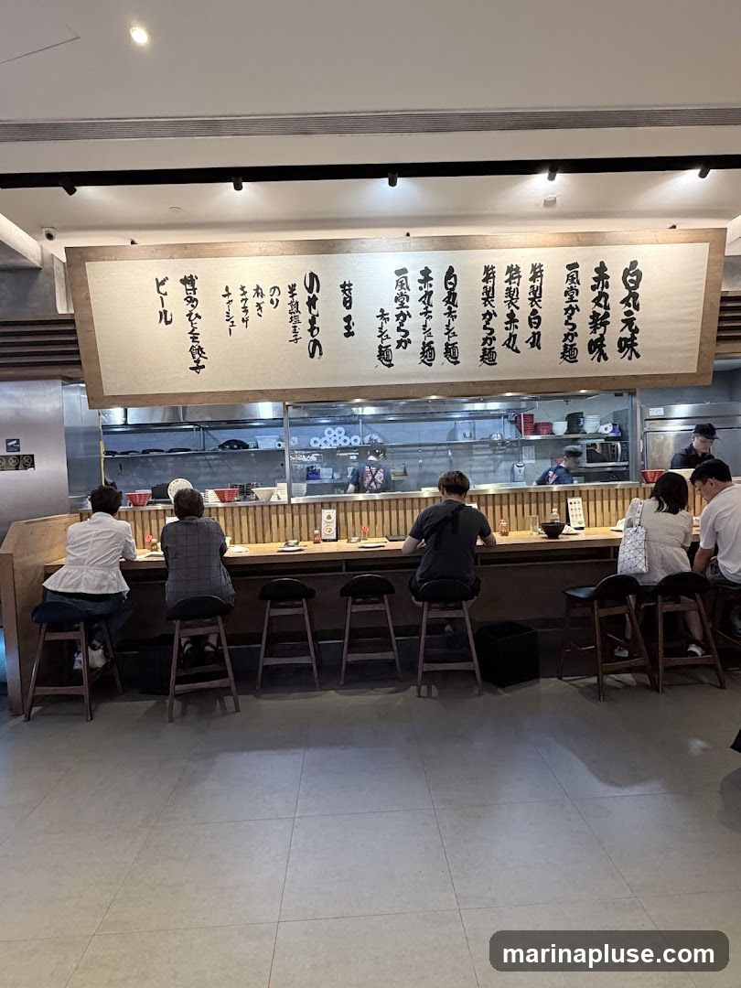 A long, horizontal menu board with Japanese text hangs above a counter where several people are seated on stools, facing the counter. The kitchen area is visible behind the counter.