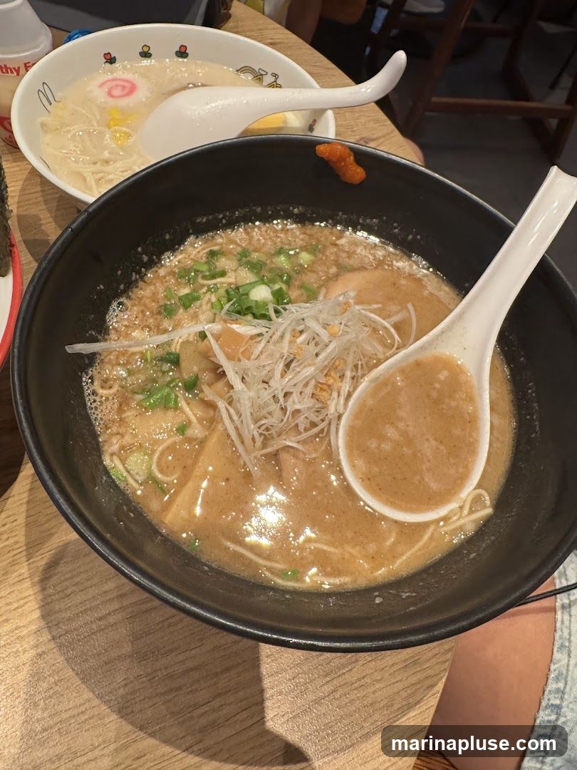 Two bowls of ramen are shown on a wooden table. The bowl in the foreground is black and filled with a rich broth, noodles, green onions, and shredded white toppings. A large white spoon rests in the broth. The bowl in the background is white and features a pink narutomaki swirl, corn, and a hard-boiled egg. A white spoon is also in this bowl. A small dollop of orange paste is on the rim of the black bowl.