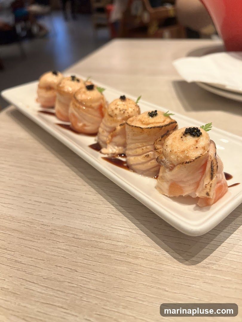 A plate of six salmon rolls topped with black caviar and a sprig of parsley, served on a white rectangular plate with a drizzle of dark sauce. The rolls appear to be seared on top. The plate is set on a light-colored wooden table. In the blurred background, other tables and patrons are visible.