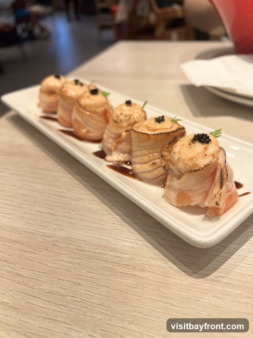 A plate of six salmon rolls topped with black caviar and a sprig of parsley, served on a white rectangular plate with a drizzle of dark sauce. The rolls appear to be seared on top. The plate is set on a light-colored wooden table. In the blurred background, other tables and patrons are visible.