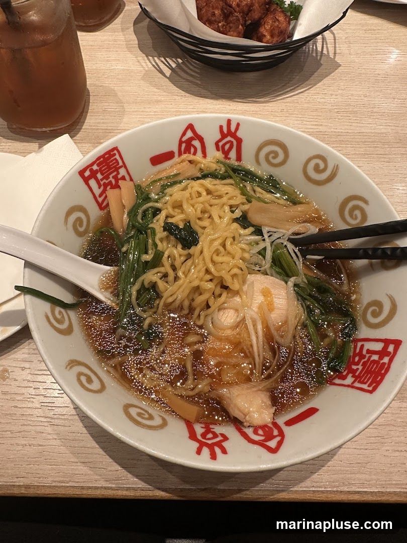 A close-up overhead view of a ramen bowl filled with noodles, broth, greens, and chicken. Chopsticks are resting on the side of the bowl. A glass of iced tea and a basket of fried food are visible in the background. The bowl has red Japanese characters and a swirl pattern on its rim.