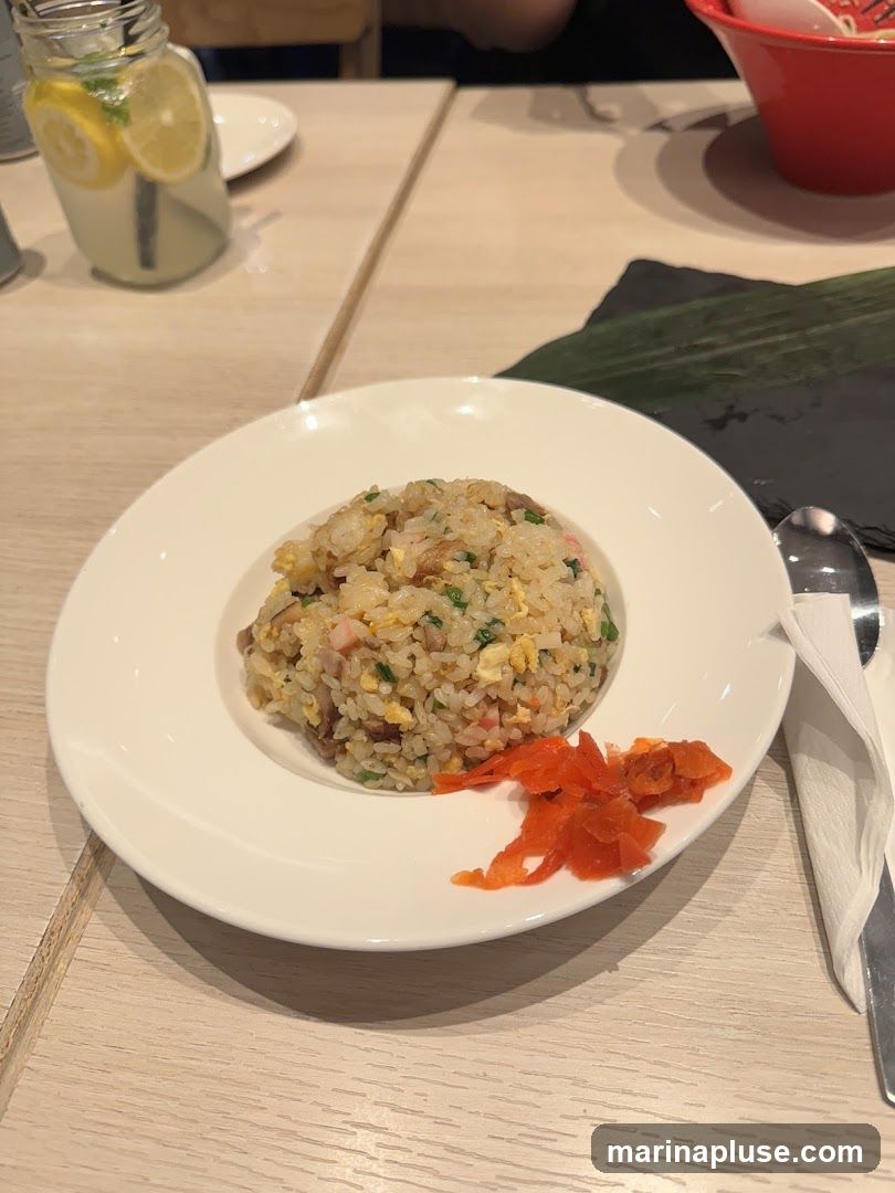 A plate of fried rice is served with pickled ginger on the side. A glass of lemonade with lemon slices and mint is visible in the background. A red bowl and a spoon are also present on the table.