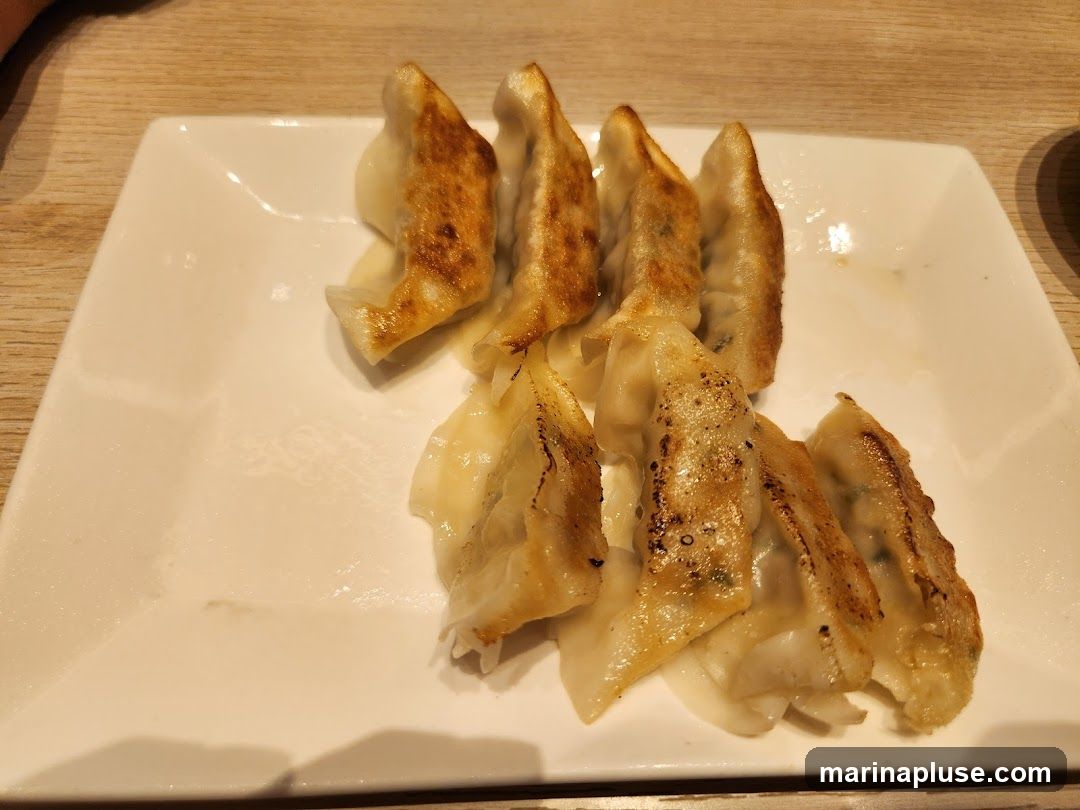 A white rectangular plate filled with pan-fried dumplings. The dumplings are golden brown on the bottom and lighter on top, with visible filling peeking through the dough. They are arranged in two rows on the plate. The table surface is visible in the background.