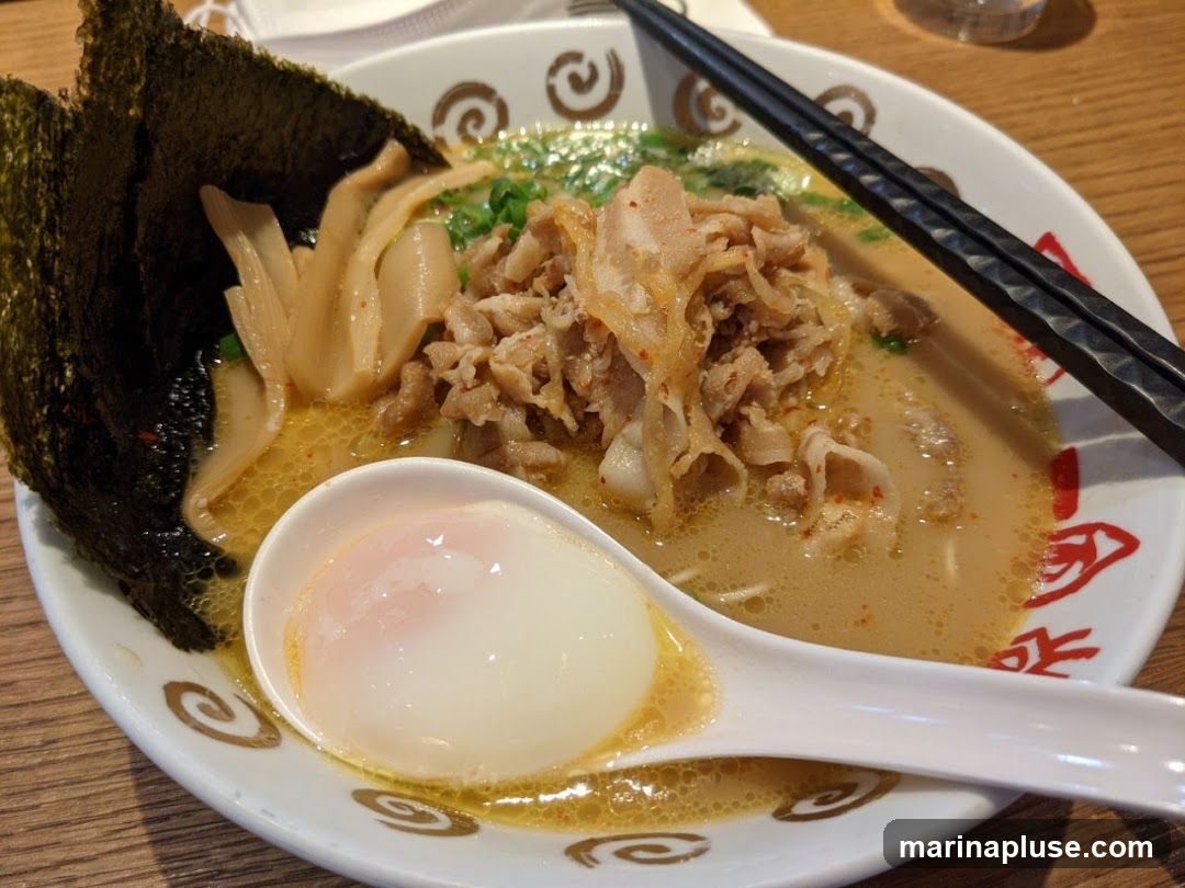 A close-up shot of a bowl of ramen. The ramen is served in a white bowl with a decorative rim. The broth is a creamy beige color, and it is topped with thinly sliced pork, bamboo shoots, and nori seaweed. A soft-boiled egg is placed in a white spoon in the foreground. Chopsticks rest on the side of the bowl.