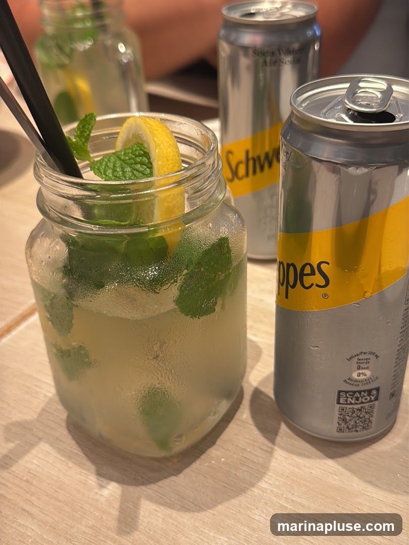 A close-up shot of a refreshing drink in a glass jar, garnished with lemon and mint. Two cans of Schweppes soda are placed next to the drink. The cans are silver with yellow branding. One can is open, and the other is unopened. The drink appears to be a mojito or a similar cocktail. The background is blurred, suggesting an indoor setting like a restaurant or bar.