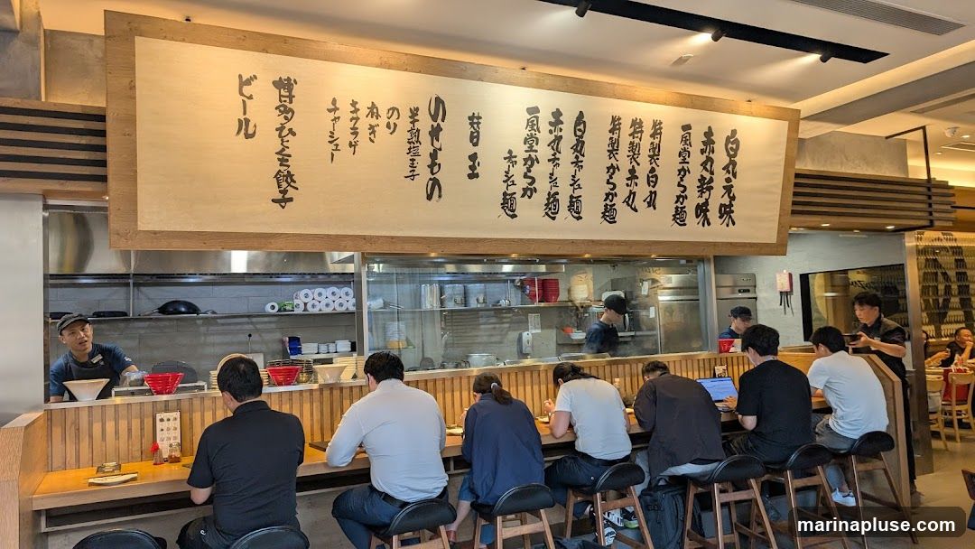A busy ramen restaurant interior with customers seated at a counter, facing a kitchen area. A large, traditional-style menu board with Japanese calligraphy hangs above the counter. Staff members are visible in the kitchen and serving customers. The decor is modern with wooden accents.