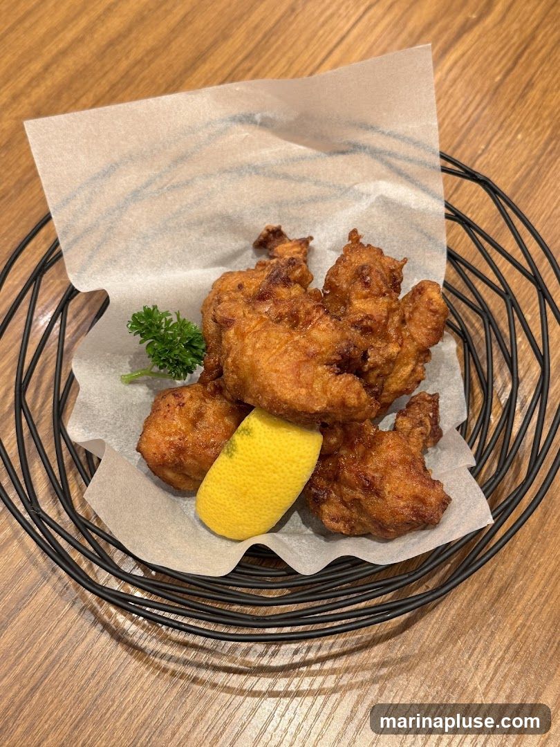 A close-up shot of several pieces of golden-brown fried chicken bites served in a black wire basket lined with white parchment paper. A wedge of lemon and a sprig of parsley garnish the dish. The food is presented on a wooden table.