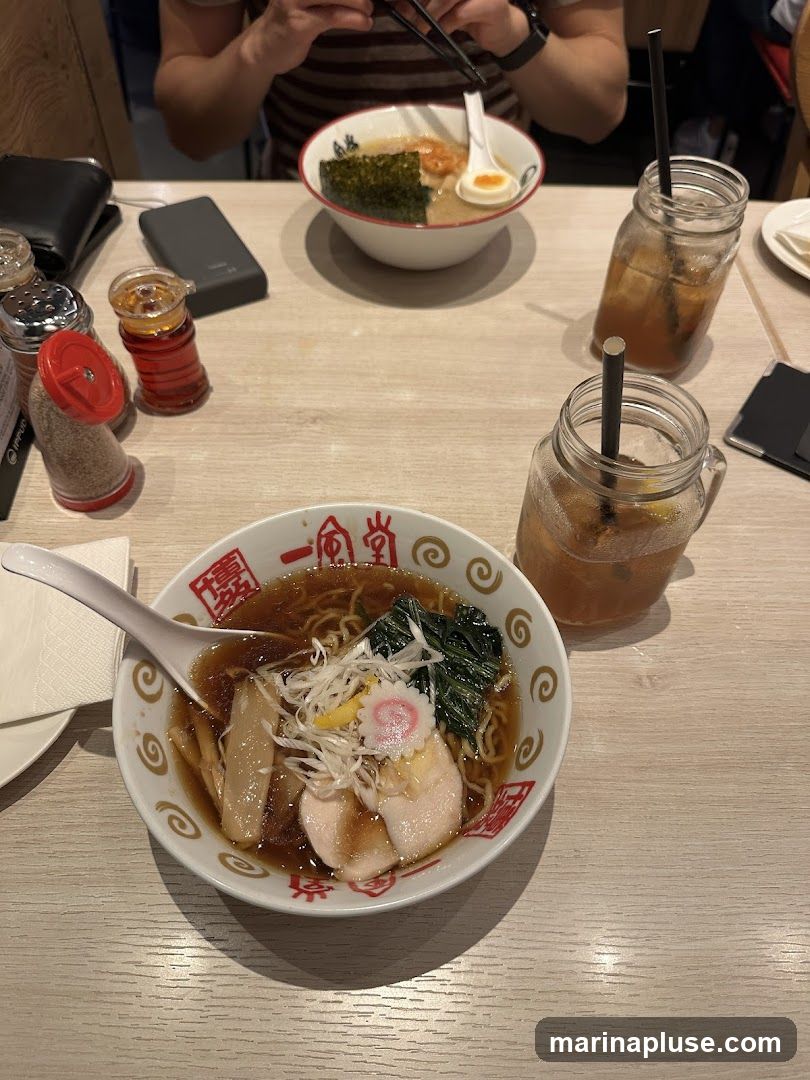 A close-up shot of two bowls of ramen and two glasses of iced tea on a wooden table. The ramen bowls are white with red Japanese characters and a swirling pattern. The bowl in the foreground contains noodles, sliced chicken, green vegetables, bamboo shoots, and a pink and white fish cake. The bowl in the background appears to have similar ingredients, including seaweed and a soft-boiled egg. The iced tea is served in mason jar-style glasses with straws. Condiment shakers and a wallet are also visible on the table.