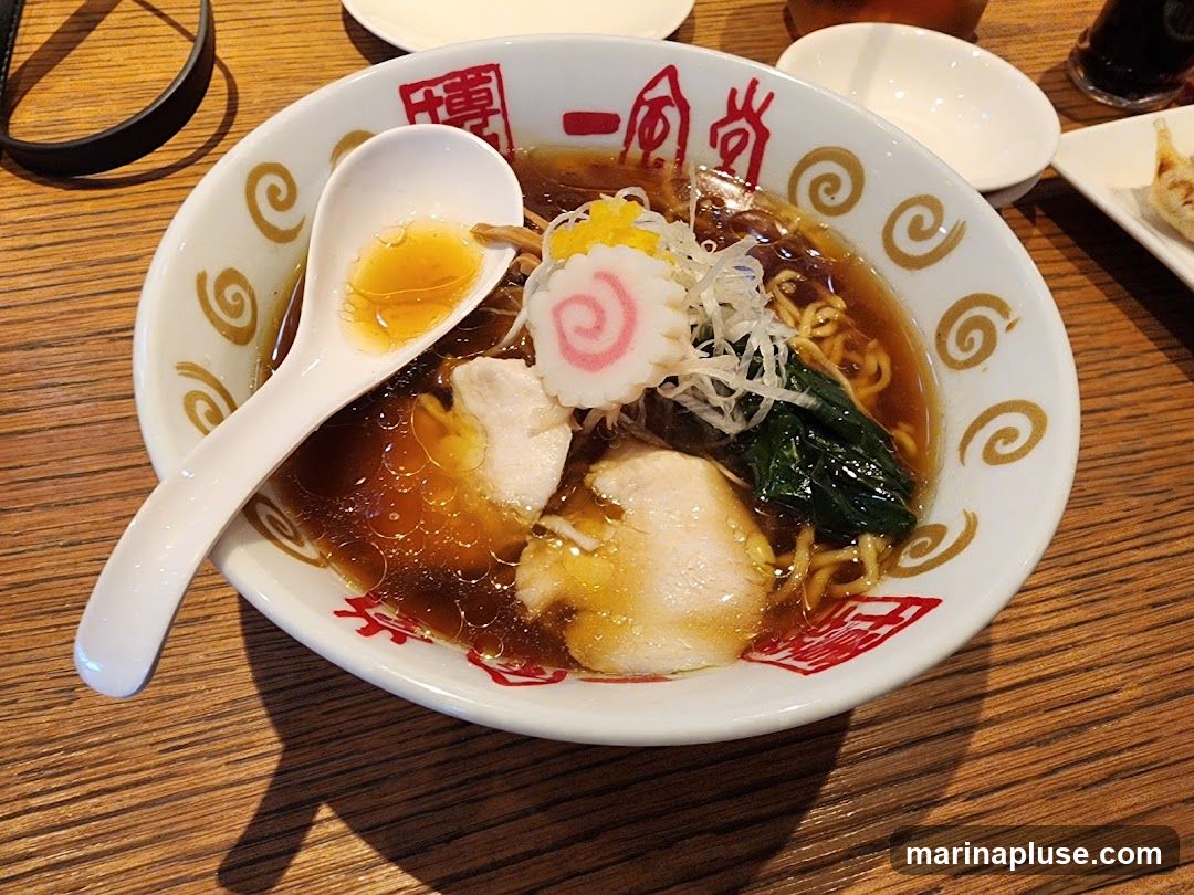 A close-up shot of a ramen bowl filled with noodles, broth, chicken, spinach, and narutomaki. A white spoon rests in the bowl, holding some broth. The bowl has a decorative rim with red Japanese characters and gold swirls. The ramen is served on a wooden table.