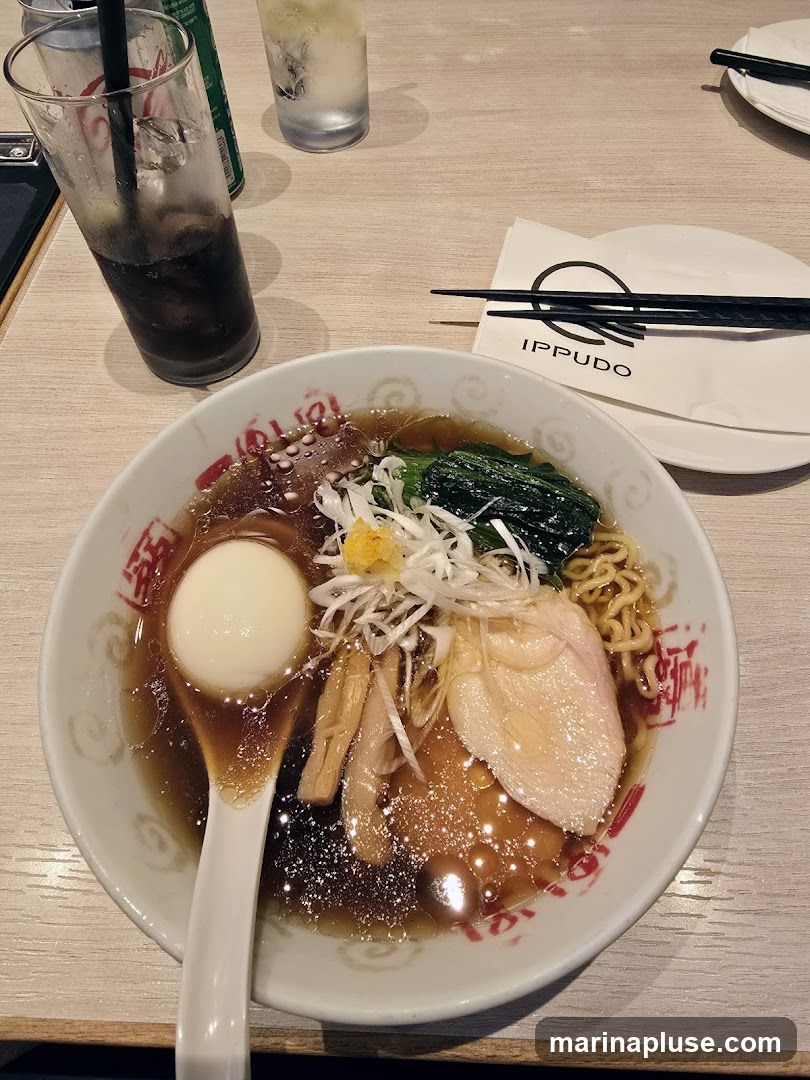 A close-up overhead shot of a bowl of ramen on a wooden table. The ramen contains noodles, broth, a soft-boiled egg, sliced chicken, bamboo shoots, and green vegetables. A white spoon rests in the bowl. To the left, a glass of dark soda with ice and a straw is visible, along with a can. To the right, a glass of water with ice and a napkin with the IPPUDO logo and chopsticks are placed on a white plate.