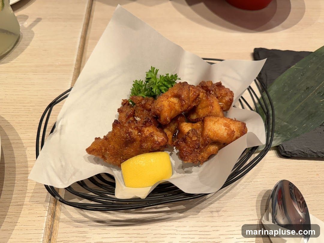 A close-up shot of several pieces of golden-brown fried chicken bites, served in a black wire basket lined with white parchment paper. A wedge of bright yellow lemon and a sprig of parsley garnish the dish. The food is placed on a light-colored wooden table. A spoon wrapped in white paper is visible in the lower right corner.