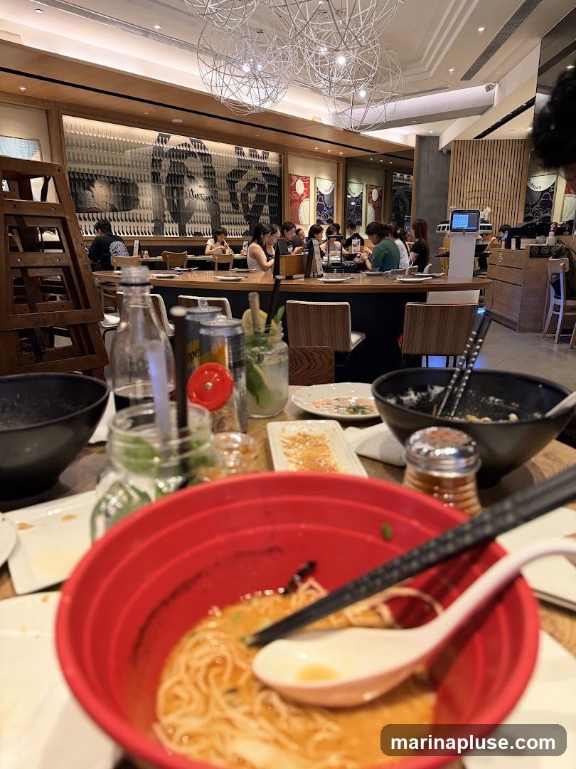 A busy ramen restaurant interior with diners seated at tables and a counter. The foreground shows partially eaten bowls of ramen, chopsticks, and drinks. The background features decorative wall art and modern light fixtures.