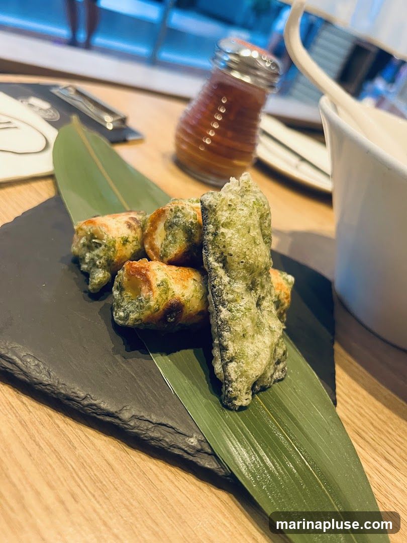 A close-up shot of a platter of fried food, possibly tempura or chikuwa, served on a dark slate with a green leaf. In the background, a glass shaker with red seasoning and a white cup with a spoon are visible on a wooden table.
