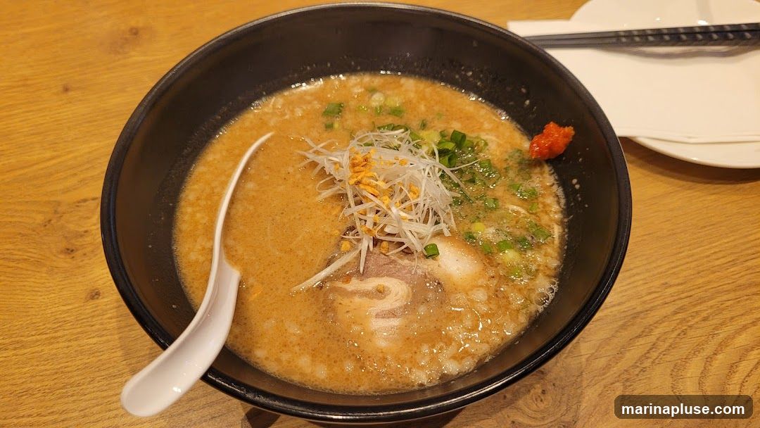 A close-up overhead shot of a bowl of ramen. The ramen has a creamy broth, topped with sliced green onions, shredded white onions, crispy garlic bits, and a slice of chashu pork. A dollop of red chili paste is visible on the side of the bowl. A white ceramic spoon rests in the broth. Chopsticks and a white plate are visible in the background on a wooden table.